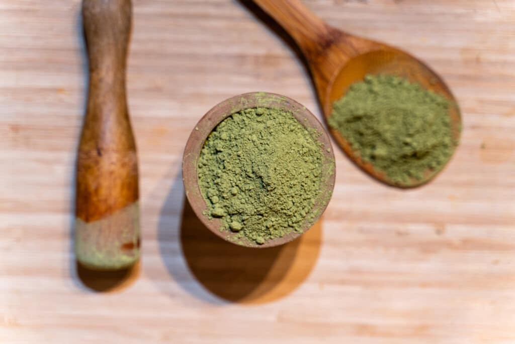 Green kratom powder in a wooden bowl with a wooden spoon beside it